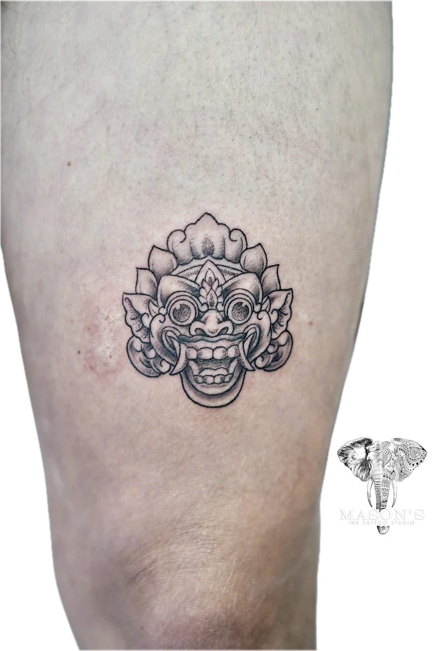 Barong Tattoo Design: a detailed ornamental face mask tattoo on the thigh