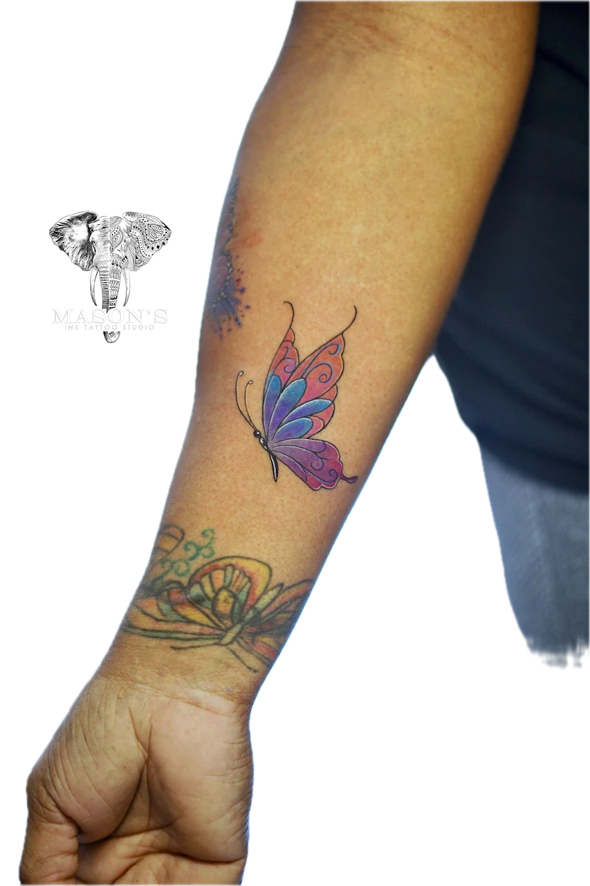 Watercolor Butterfly Tattoo by Mason’s Ink: a colorful butterfly and flower with vines on a forearm
